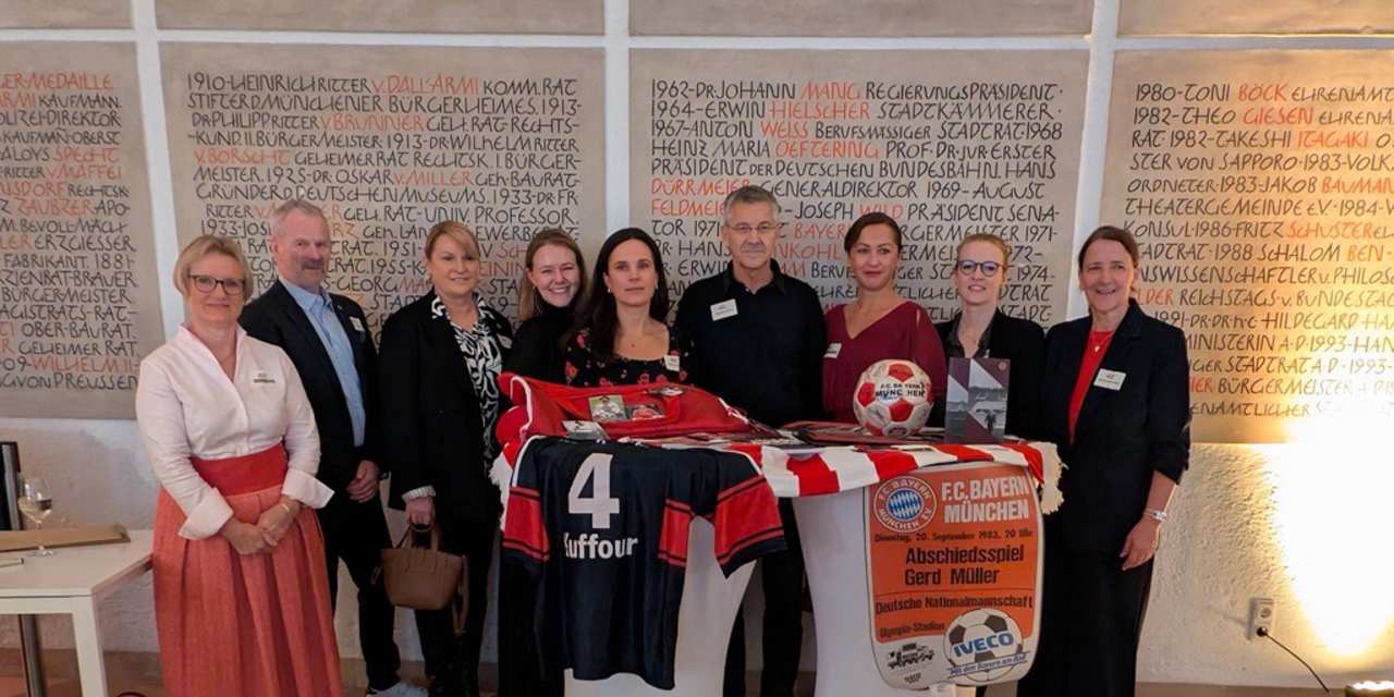 Herbert Hainer steht mit Verantwortlichen zum Jubiläum der Alzheimergesellschaft an einem Stehtisch und zeigt den Erinnerungskoffer des FC Bayern