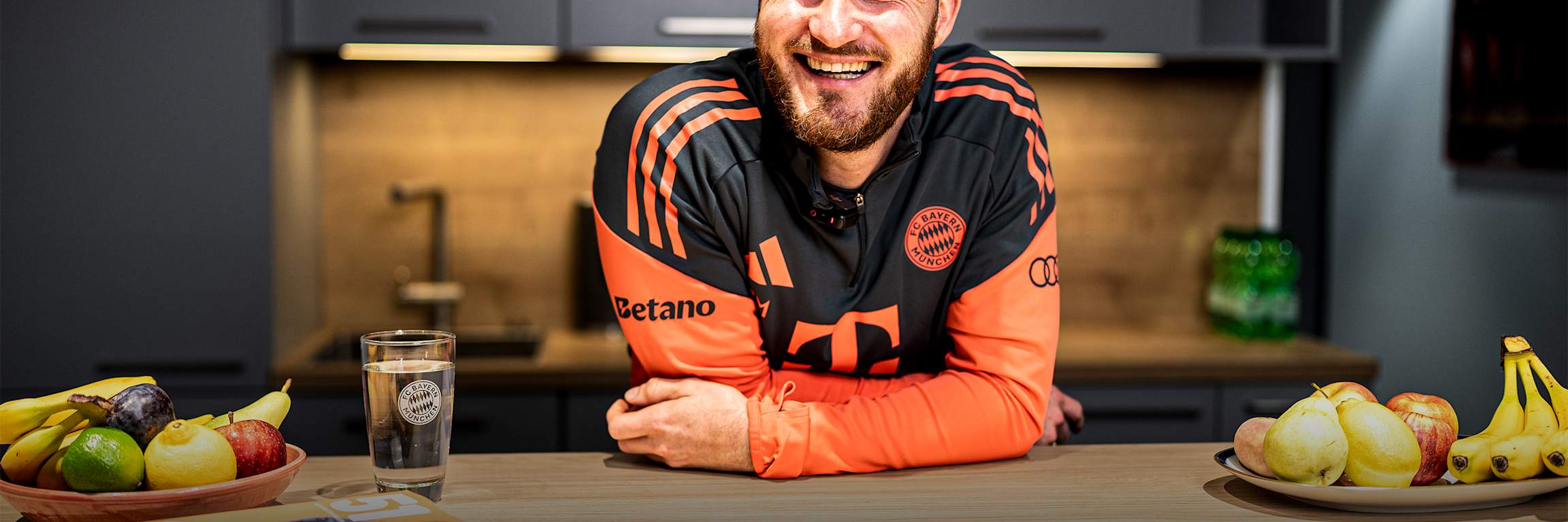 Melf Carstensen lehnt in der FC Bayern Teamline lachend über einen Tisch.