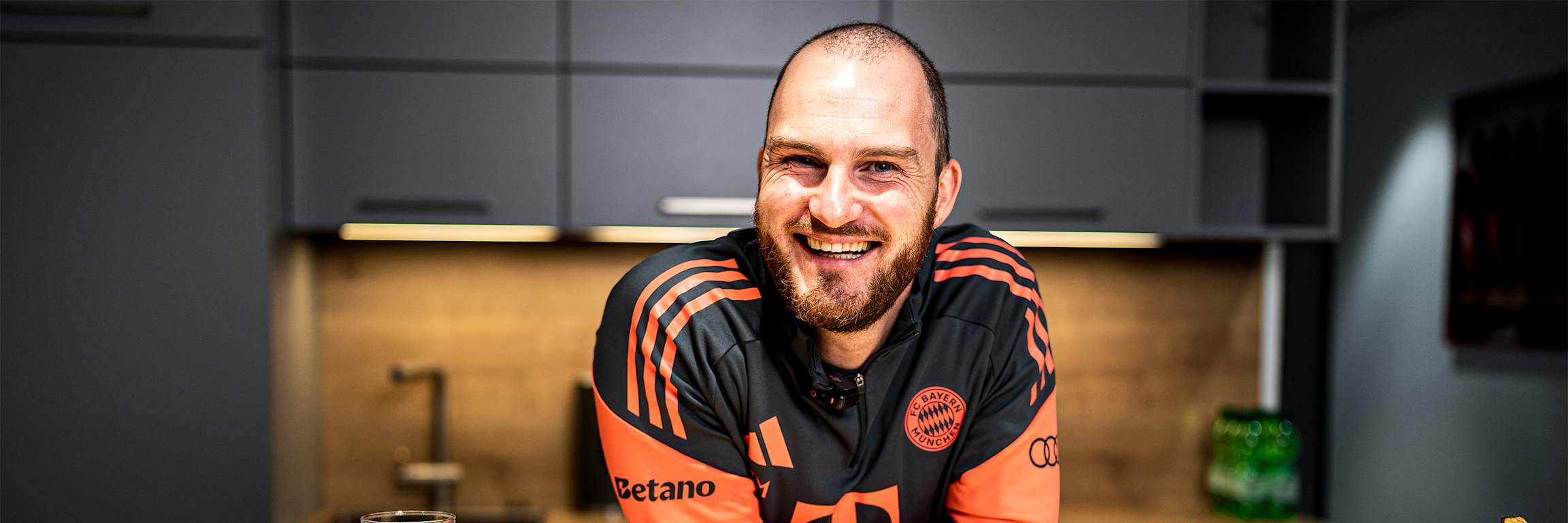 Melf Carstensen lehnt in der FC Bayern Teamline lachend über einen Tisch.