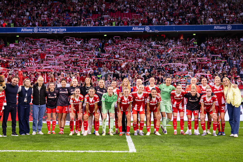 Die FC Bayern Frauen in einem Gruppenfoto vor der Südkurve der Allianz Arena mit Fans im Hintergrund.