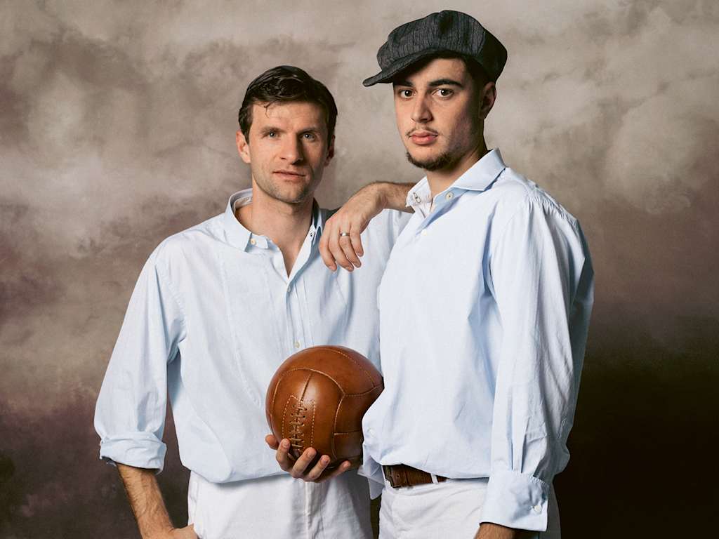 Thomas Müller und Aleks Pavlovic stehen nebeneinander in alter Spielkleidung des FC Bayern, Pavlovic hält einen Lederball in der Hand