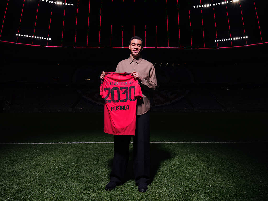 Jamal Musiala steht auf dem Rasen der Allianz Arena und hält ein Trikot mit 2030 und Musiala hoch