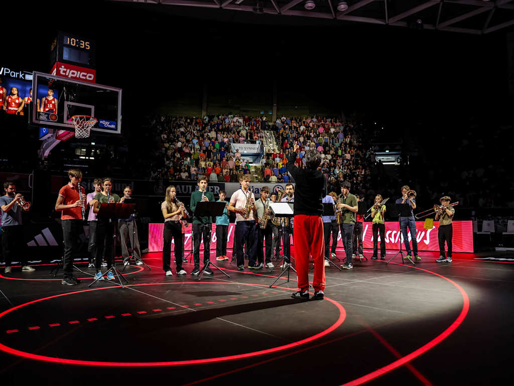 Ein Orchester von Jugendlichen spielt auf der Basketball-Spielfläche, auf der Tribüne dahinter steht der Chor der Jugendlichen und singt.