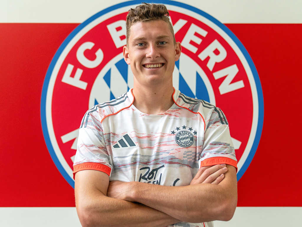 David Heindl und posiert im Trikot vor dem FC Bayern-Wappen am FC Bayern Campus.
