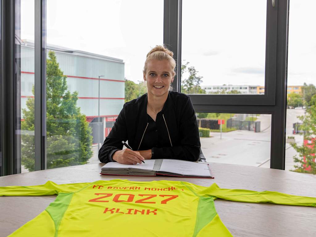 Anna Klink sitzt am Tisch und unterschreibt ihren Vertrag. Sie lächelt in die Kamera, vor ihr liegt ihr Trikot mit der Laufzeit 2027.