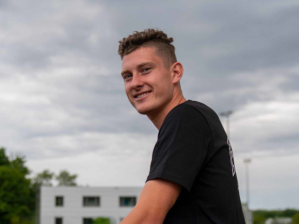 Alt-Tag: David Heindl steht mit verschränkten Armen und einem Lächeln vor dem FC Bayern Logo