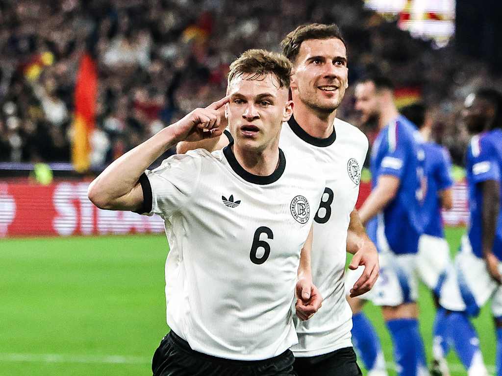 Joshua Kimmich jubelt im Trikot der deutschen Nationalmannschaft und zeigt mit dem Finger auf seine Stirn. Dahinter läuft Leon Goretzka.