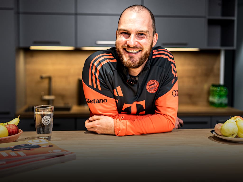 Melf Carstensen leans on a table smiling wearing the FC Bayern Teamline gear