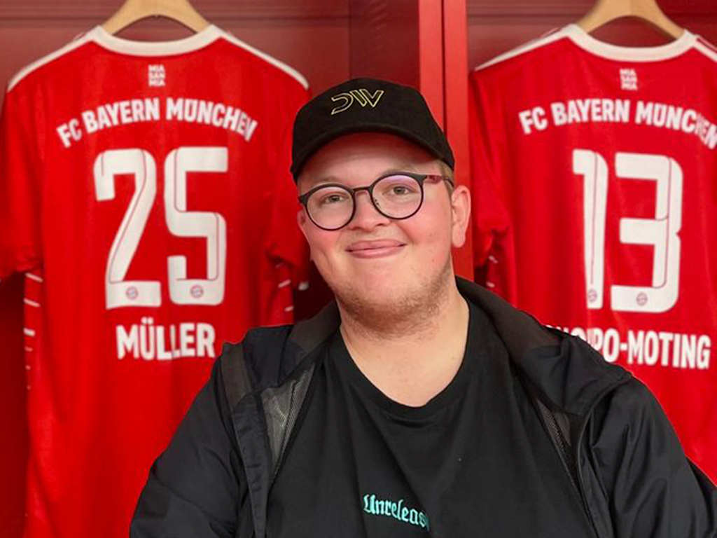 FC Bayern-Fan Glenn sitzt in der Kabine der Allianz Arena vor einem Trikot von Thomas Müller