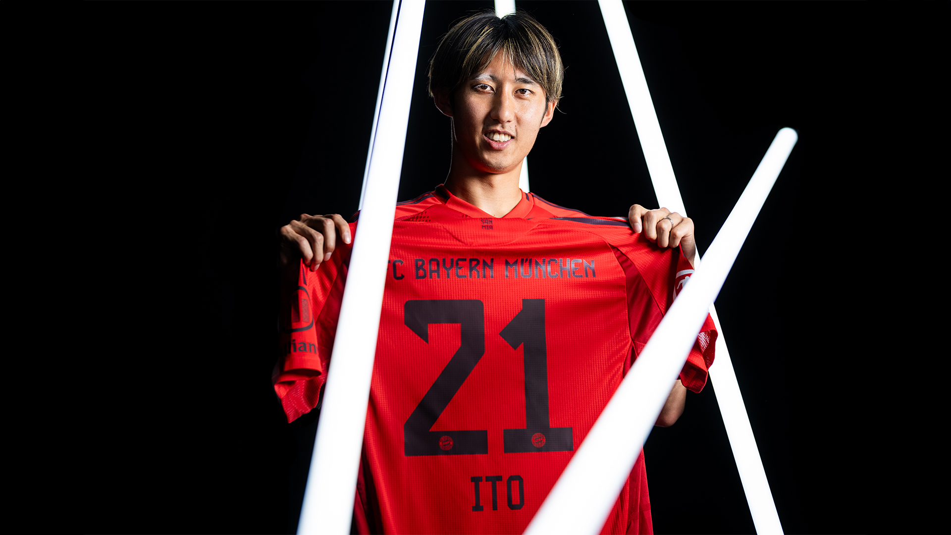 Hiroki Ito hält das Trikot mit der Nummer 21 hoch, in dem er zukünftig für den FC Bayern auflaufen wird.