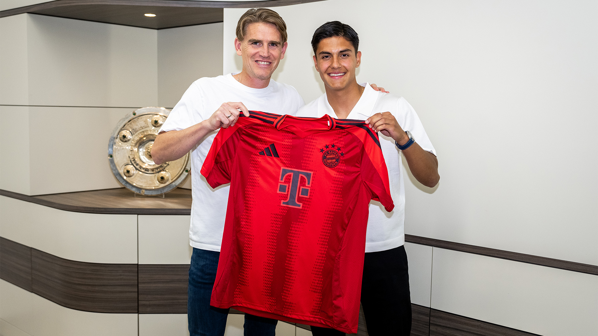 Christoph Freund und Matteo Perez Vinlöf halten einen Trikot hoch, nachdem dieser einen Profivertrag beim FC Bayern unterschrieben hat.