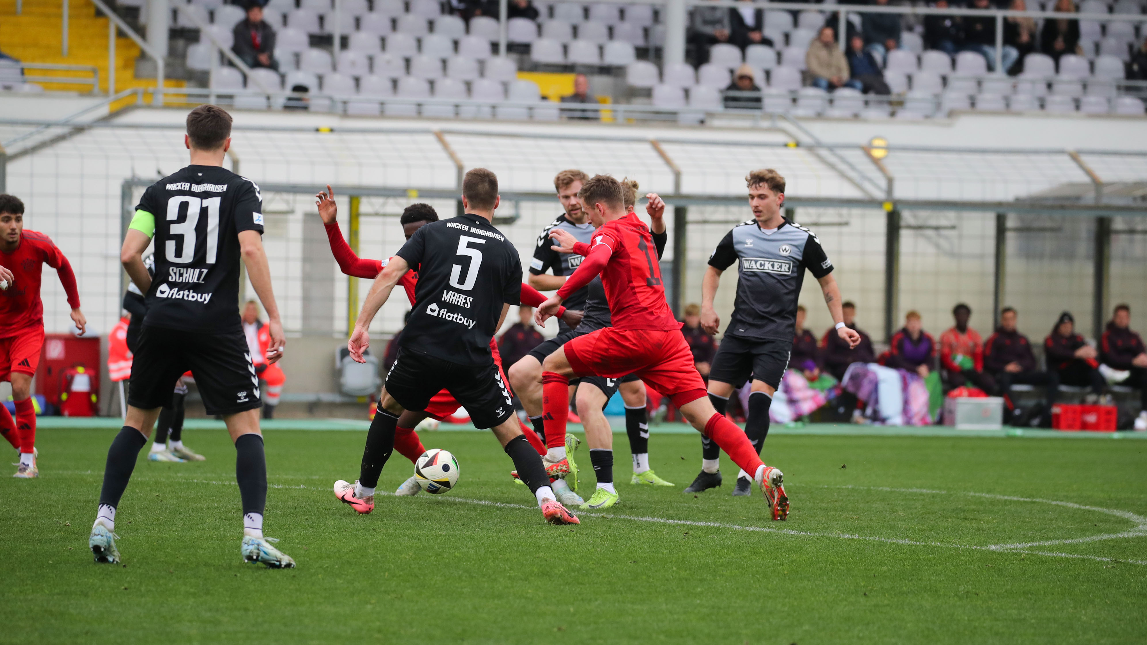 Timo Kern von den FC Bayern Amateuren im roten Trikot im Zweikampf mit Wacker Burghausen-Spielern in Schwarz während eines Fußballspiels in der Regionalliga.